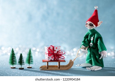 Cute Cheerful Santas Helper Elf Pulling Santas Sleigh With Christmas Present. North Pole Christmas Scene. Santas Workshop. Elf At Work. 