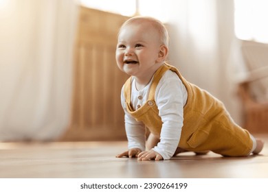 Cute cheerful smiling blonde little baby boy discovering world, crawling on wooden floor by home, copy space. Positive happy infant child playing in living room interior