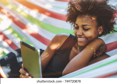 Cute cheerful teenage Brazilian female with African hair is laying in colorful striped hammock located in public park on warm sunny day and smiling while reading funny memes on her digital tablet
