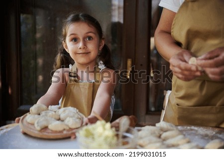 Cute child, beautiful Caucasian little girl wearing a beige chef's apron, smiling a cheerful sweet smile, holding a wooden board with homemade dumplings. Artisanal, rural scene of cooking food