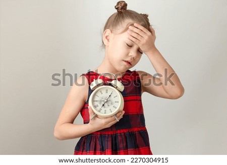 Cute child with closed eyes and hand on head holding alarm clock concept to wake up early, caucasian kid little girl of 6 7 years in red plaid dress