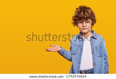 Cute child in denim shirt and glasses looking at camera and pointing at blank space isolated on yellow background