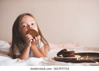 Cute Child Girl 3-4 Year Old Lying In Bed Eating Chocolate Cookies Over Glow Christmas Lights Wear Pajamas Close Up. Childhood. Healthy Lifestyle. 