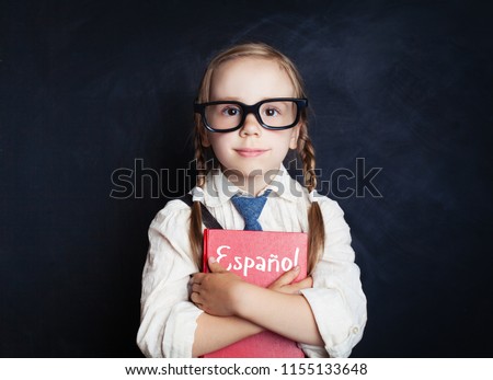 Cute child girl embracing spanish book against chalkboard background in school classroom. Learning spanish and education concept
