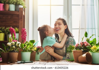 Cute Child Girl Helps Her Mother To Care For Plants. Mom And Her Daughter Engaged In Gardening Near Window At Home. Happy Family In Spring Day.