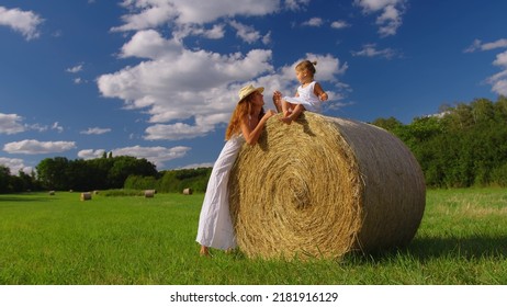 Cute Child Girl And Her Mother In White Dresses Smile In The Green Meadow. Mother And Child Enjoy With Beautiful Nature And Sun Rays At Sunset Time Together In Slow Motion Shot