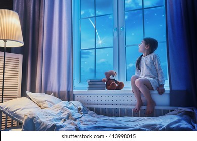 Cute child girl sitting at the window and looking at the stars. Girl making a wish by seeing a shooting star at bedtime night.