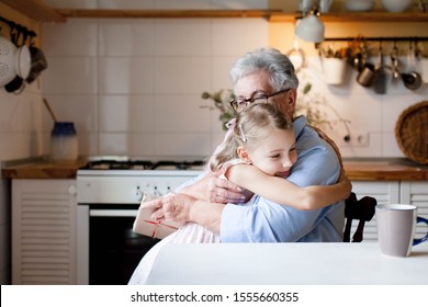 Cute Child Is Giving Gift Box To Grandmother. Kid Girl And Senior Woman Are Hugging In Cozy Kitchen At Home. Happy Family Is Enjoying Holidays, Kindness, Togetherness. Lifestyle Authentic Moment.