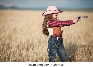 Cute Child Having Fun Outdoors, Little Girl Cowboy Playing In Wheat Field At Sunset. Happy Baby Girl With Toy Gun And Cowboy Hat Enjoying Nature. American Cowgirl. Lovely Smiling Toddler Portrait.