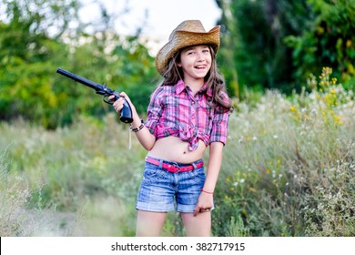 Cute Child Having Fun Outdoors, Little Girl Cowboy Playing In Wheat Field At Sunset. Happy Baby Girl With Toy Gun And Cowboy Hat Enjoying Nature. American Cowgirl. Lovely Smiling Toddler Portrait.