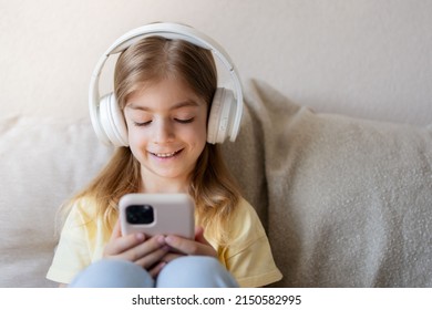 Cute Child With Headphones And Mobile Phone On Sofa Indoors.