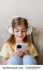 Cute Child With Headphones And Mobile Phone On Sofa Indoors.