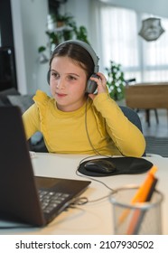 Cute Child With Headphones Using Laptop Computer For Online Learning. Home School. Girl Doing Homework At Home. Lifestyle Concept For Home Schooling. Social Distancing And Education.