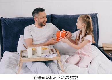 Cute Child Presenting Fathers Day Gift Box To Daddy Having Breakfast In Bed