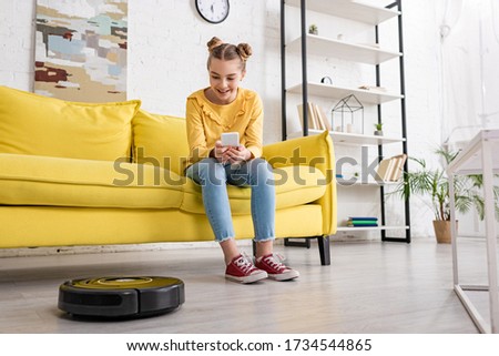 Cute child with smartphone on sofa smiling and looking at robotic vacuum cleaner on floor in living room Stock photo © 