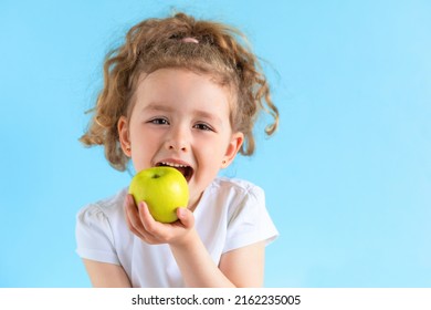 Cute Child Smiling Bites Apple. Nutrition For Caries Prevention. Portrait Of Little Girl With Fruit On Blue Background. Kid Eating Healthy Food, Snack. Teeth, Dental Health, Smile. Children Dentistry