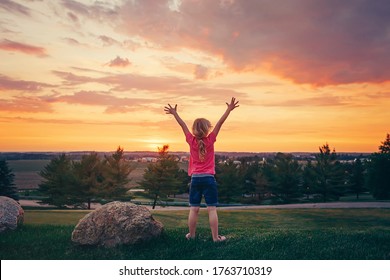 Cute Child Standing With Her Arms Raised On Hill Outdoor At Sunset.  Girl Kid Having Fun Outdoor Enjoying Life At Sunrise. Local Staycation Vacation Travel. View From Back. Happy Childhood Lifestyle.