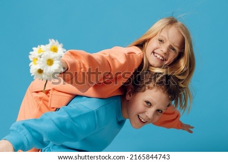 cute children, brother and sister stand sideways to the camera and the fool, the boy rolls the girl with a bouquet of daisies in his hand on his back, and she joyfully holds out her hand to the camera