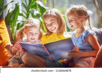 Cute children girls are reading a book sitting on sofa in room at home.