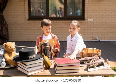 Cute Children Standing At Table With Various Goods And Buying Toy And Book At Garage Sale