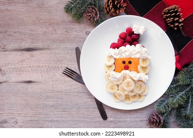 Cute Christmas Santa Claus Pancake On A White Plate With Raspberry Hat And Banana Beard. Top Down Table Scene Against A Wood Background.