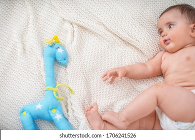 Cute Chubby Baby 4 Months Old Is Lying On His Side On Bed With Toy. Child Pulls Handles To A Toy Giraffe Made Of Fabric. The Baby Learns To Roll Over On Her Tummy. Copy Space. Kid Learns The World.