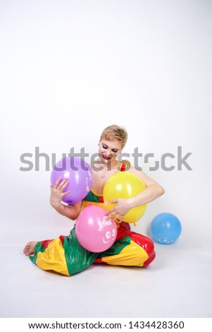 cute chubby girl with short blonde hair and plus size figure in bright colored PVC jumpsuit posing with air balloons on white background alone. playful young woman in halloween good clown costume.