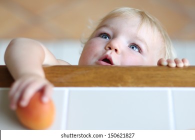 Cute Clever Baby With Beautiful Blue Eyes And Blond Hair Standing On Tiptoes Reaching Out Tasty Apricot From High Wooden Table