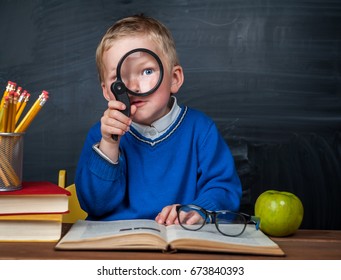 Cute Clever Boy Is Sitting At A Desk With Magnifying Glass In Hand. Child Is Reading A Book With A Blackboard On A Background. Ready For School. Back To School. Apple And Books On Desk.