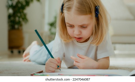 Cute concentrated Caucasian little kid girl draw color pencil lying floor at home play alone  Talented preschool child daughter kid baby coloring picture at home drawing painting creative paint