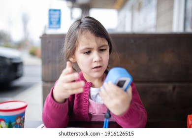 Cute And Concerned-looking Little Girl In Pink Sweater Holding Small Camera For Kids. Adorable Young Child Playing With Toy Camera With Blurry Background. Kids Interacting With Technology