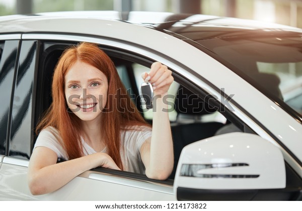 Cute content red haired housewife in a white car\
holding car key in her hand and looking at camera. Car Rent or Auto\
purchase Concept.