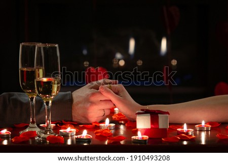 Cute couple celebrating Valentine’s Day, holding hands lovingly during romantic dinner. Red roses, gift, candles and glasses of champagne. Concept about lifestyle, people and celebrations.