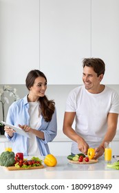 Cute Couple Cooking In Kitchen, Man Chopping Vegetables While His Girlfriend Is Using A Digital Tablet Looking For A Recipe. Family Enjoying Weekend Together.