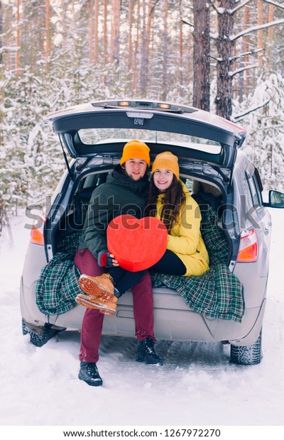 cute couple in love with big red heart\
sitting in a car with an open trunk and kissing in the winter\
forest  between  of the trees covered with\
snow