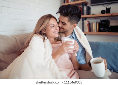 Cute Couple Relaxing On Couch Under Blanket At Home In The Living Room And Drink Coffee.