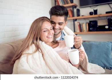 Cute Couple Relaxing On Couch Under Blanket At Home In The Living Room And Drink Coffee.