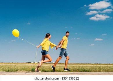 Cute couple running and holding hands. In background field. Girl holding yellow balloon. Dominating blue and yellow colors.