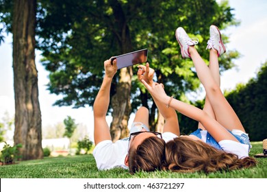 Cute Couple Of Young People Is Lying On Grass In Summer Park. Guy In White T-shirt Holds Tablet Overhead, Girl With Long Curly Hair Holds Legs Up.