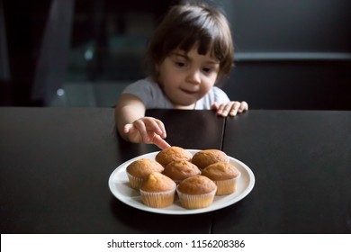 Cute Curious Little Girl Looking And Reaching Hand Taking Muffins On Table, Hungry Funny Kid Eager To Eat Cake Stealing Cookie While Parents Not Watching, Bakery And Child Addiction To Sweets Concept