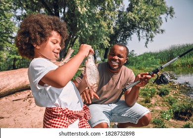 Cute curly Afro-American boy looking at the fish in his hands while his dad holding a fishing rod - Powered by Shutterstock