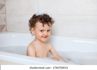 Cute Curly Baby Bathes In The Bath With An Emotional Laugh. Personal Hygiene Rules For Children. Boy, A Child Plays With Toys In A Full Bath With Water