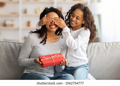 Cute curly black girl making birthday surprise for her excited mom, home interior, copy space. Happy african american daughter closing her mother eyes while sitting on couch, woman holding gift box