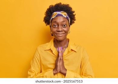 Cute Curly Haired Afro American Woman With Dark Skin Keeps Palms Pressed Together Asks For Favor Smiles Gently Wears Shirt And Headband Isolated Over Yellow Vivid Background. Thank You Very Much