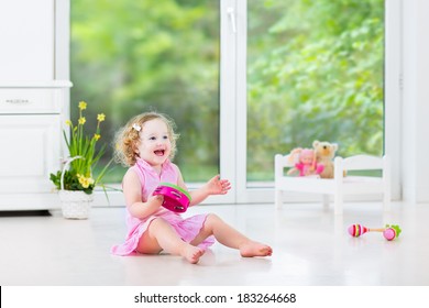 Cute Curly Laughing Toddler Girl In A Pink Dress Playing Tambourine And Maracas In A Sunny Room With A Big Garden View Window With A Toy Bed And Spring Flowers Next To Her, White Modern Interior