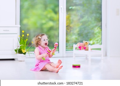 Cute Curly Laughing Toddler Girl In A Pink Dress Playing Tambourine And Maracas In A Sunny Room With A Big Garden View Window With A Toy Bed And Spring Flowers Next To Her, White Modern Interior Home