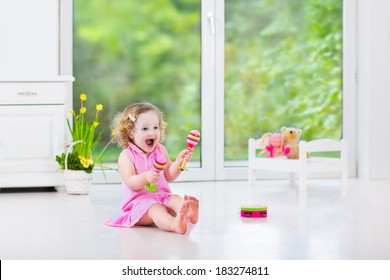 Cute Curly Laughing Toddler Girl In A Pink Dress Playing Tambourine And Maracas In A Sunny Room With A Big Garden View Window With A Toy Bed And Spring Flowers Next To Her, White Modern Interior Home