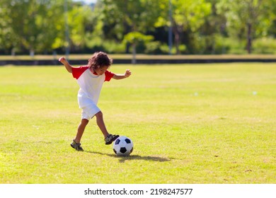 Cute Curly Little Boy Playing Football. Kids Play On Outdoor Pitch. Adorable Little Athlete Kicking Ball. Summer Outdoor Fun. Toddler Running After Ball. Sports For Active Child. Children Exercise.