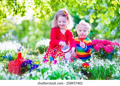 Cute Curly Little Girl In A Red Summer Dress And Funny Baby Boy Working In The Garden Watering First Spring Flowers On A Sunny Warm Day. Kids Gardening. Children Having Fun Together Outdoors.