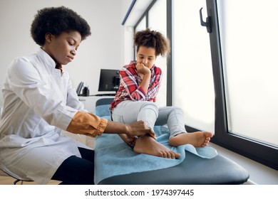 Cute curly mixed race teen 12-aged girl, sitting on the couch at modern clinic, while young 30-aged female African doctor orthopedist rheumatologist examining patient's leg - Powered by Shutterstock